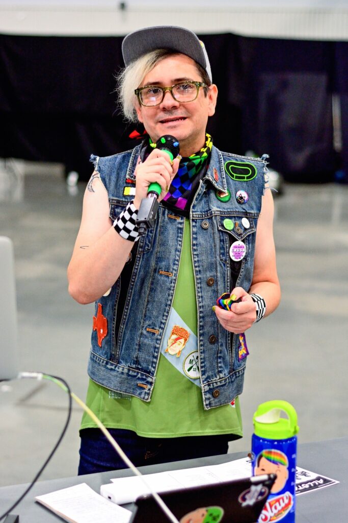Thin Mint stands indoors holding a microphone and smiling while announcing at a roller derby event. They wear a gray cap, glasses, a sleeveless denim vest covered in colorful patches and pins, a green shirt, and a checkered wristband. A rainbow-patterned bandana is around their neck, and they hold a small rainbow fidget cube in their other hand. In front of them is a table with papers, an iPad covered in stickers, and a bright green water bottle. The background is blurred, highlighting them as the focal point.